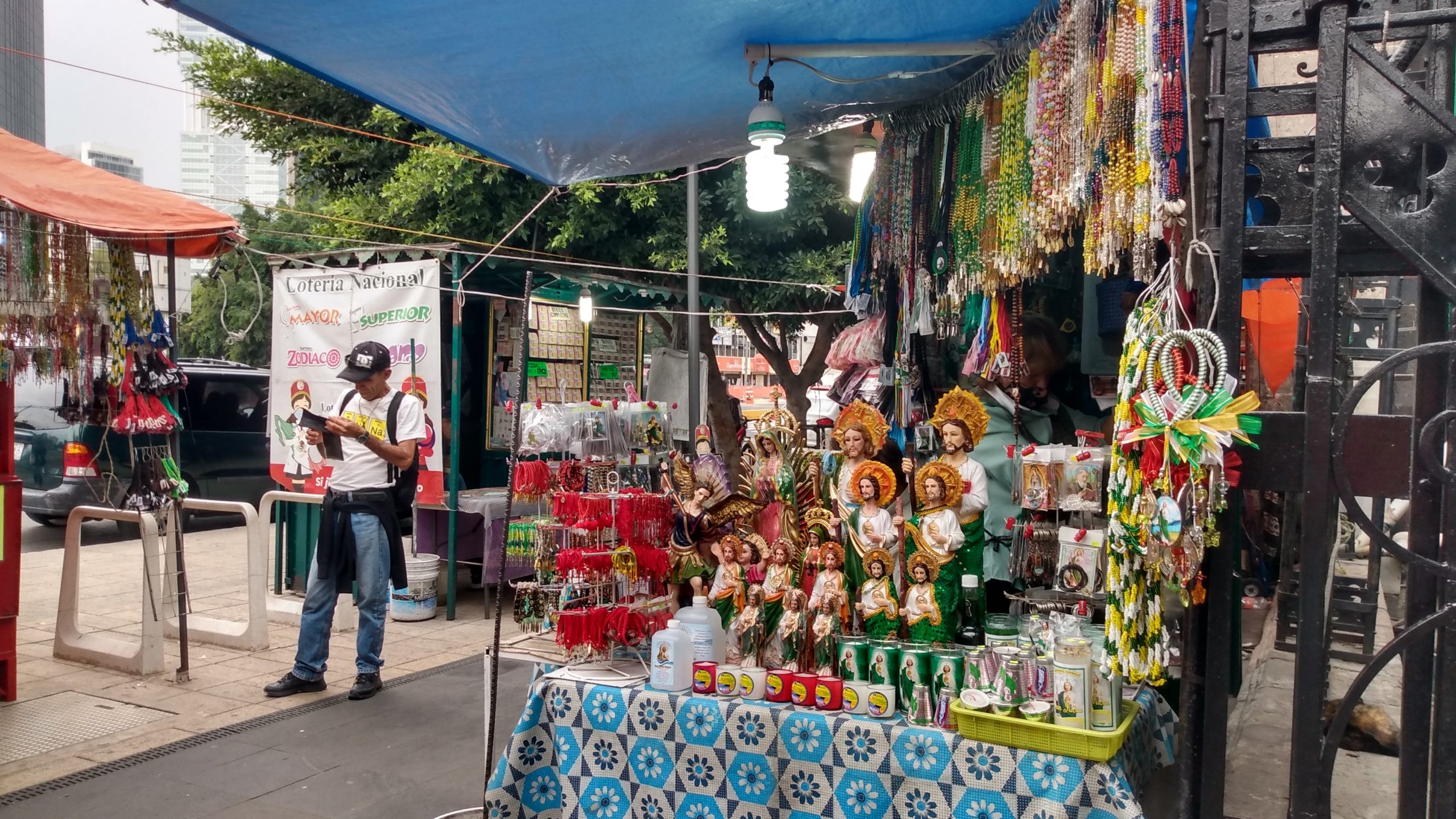 Un largo puesto de collares, pulseras, veladoras y de más objetos de San Judas Tadeo. A la izquierda, uno de los muros de la Iglesia de San Hipólito. | Foto: Diana Hernández / Conexión Migrante.