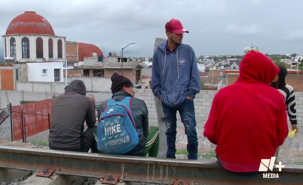 Migrantes en Apizaco en las vías del tren, con la barda metálica de fondo.